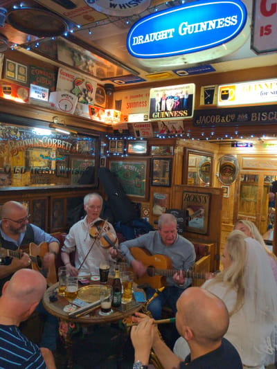 The image depicts an interior scene of what appears to be a traditional Irish pub. The walls are adorned with numerous vintage signs advertising brands such as Guinness, Old Irish Whiskey, and John Jameson. Wooden paneling and furniture contribute to the rustic ambiance. In the foreground, a group of people are gathered around a circular table. A man on the left is playing an acoustic guitar. To his right, another man is playing the violin. A woman with blonde hair, possibly wearing a veil, is seated on the right. There are drinks on the table, including glasses of beer and a bottle. The overall impression is of a lively gathering, possibly a musical performance, within a historic establishment. The mention of Belfast, United Kingdom, is not directly evident from the visual information within the image itself, but the presence of Irish-themed advertisements and the general aesthetic strongly suggest a location within Ireland, and Belfast is a plausible setting for such a pub.