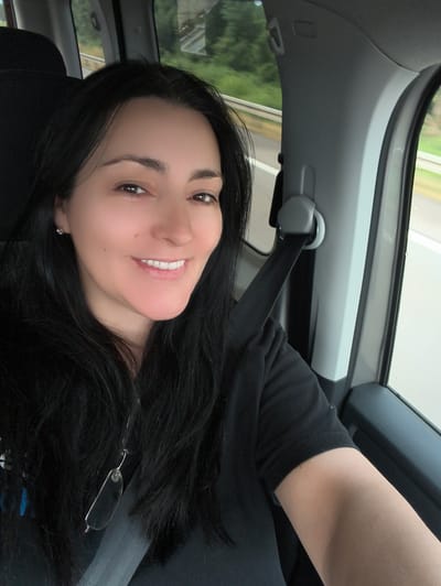 A dark-haired woman with fair skin is smiling for a selfie inside a vehicle. She is wearing a black t-shirt and a silver earring in her left ear. A pair of eyeglasses is hanging from her neck by a thin string. The woman is seated in the passenger seat, and the seatbelt is visible across her chest. The interior of the car, including the gray pillar and window frame, is visible. Outside the window, a green, tree-lined landscape and a road can be seen, suggesting the vehicle is in motion. The background scenery is blurred due to movement. The image is taken from a slightly elevated angle.