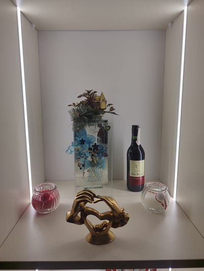 This image depicts a shelf display inside a retail establishment in Chuy, Kyrgyzstan. The shelf is illuminated by vertical LED light strips on either side. On the shelf, from left to right, are a red candle in a fluted glass jar, a gold-colored sculpture of two hands forming a heart with a small figure inside, a tall clear glass vase containing a blue and gold floral arrangement with greenery, a bottle of red wine labeled "Omar Khayyam VIN ROUGE," and a clear glass jar containing a white pom-pom tied with red and white string. The background is a plain white wall.