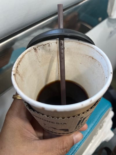 A close-up, eye-level, outdoor shot shows a hand holding a disposable paper coffee cup with a dark lid and brown straw. The cup has a cardboard sleeve that reads "INDONESIA". The cup contains a small amount of dark liquid. The background is blurred, showing a reflective surface and some teal and white material. There are no visible signs of the specific location within Dukuhturi, Indonesia.