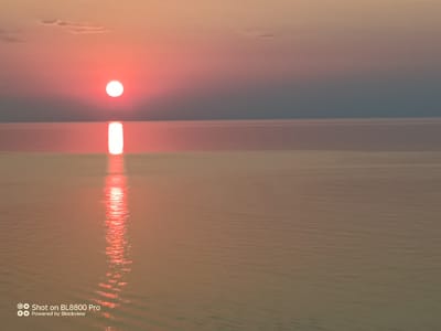 The image depicts a serene seascape with a vibrant sunset over the Black Sea, near Kamen Bryag, Bulgaria. The sun, a bright orange orb, is positioned in the upper left quadrant of the sky, casting a long, shimmering reflection on the calm water. The sky transitions from a soft pink and orange near the horizon to a dusky grey at the top. Wisps of clouds are faintly visible against the lighter part of the sky. The water surface is smooth, with gentle ripples catching the light, creating a path of golden luminescence leading from the sun towards the foreground. The overall atmosphere is tranquil and picturesque, characteristic of the coastal beauty found in this region of Bulgaria.