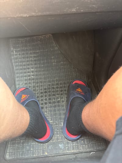 This image shows a pair of feet wearing navy blue Superman-themed slippers over black socks, resting on a black rubber car floor mat. The mat has a grid pattern. The background appears to be the interior of a vehicle, with dark grey plastic surfaces visible. The lighting suggests it is daytime. The location is specified as Mikorzyn, Poland.