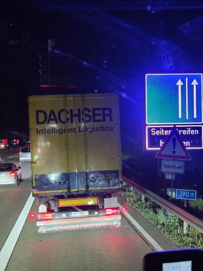 The image depicts a nighttime view from a vehicle on a highway, likely in or near Obertshausen, Germany, given the presence of German text on signs and the truck. In the foreground, the rear of a large yellow semi-trailer truck is visible. The truck displays the logo "DACHSER Intelligent Logistics" in dark blue lettering. The license plate of the truck is partially visible and reads "8H7*0928". On the rear of the truck, there are brake lights and a speed limit sign indicating "90 km".

To the right of the truck, a digital road sign is illuminated with blue light. The top panel of this sign shows two vertical arrows pointing upwards, indicating multiple lanes are open. The panel below it reads "Seitenreifen" and "be" and "en", with a triangular warning sign featuring an exclamation mark, suggesting caution due to side conditions or obstacles. Below this, another sign reads "Spurrinne", which translates to "rut" or "groove" in the road, further emphasizing the warning. A blue rectangular sign displays "190.0", likely indicating a distance or a point marker.

The highway appears to be congested, with other vehicles visible in the distance. A white van and a white car are seen ahead of the truck. The road is marked with white lines, and there is a grassy verge and a guardrail to the right of the highway. The overall environment is dark, characteristic of nighttime driving on a highway.