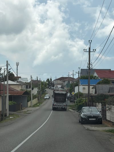 The image captures a street scene in Sergey-Pole, Russia, under a cloudy sky. A garbage truck is traveling down a paved road lined with houses and utility poles. To the right, a black BMW is parked on the side of the road. Several other cars are visible further down the street. The environment appears to be a residential area with a mix of residential buildings and some commercial or industrial structures. The presence of utility poles and overhead wires suggests an established infrastructure.