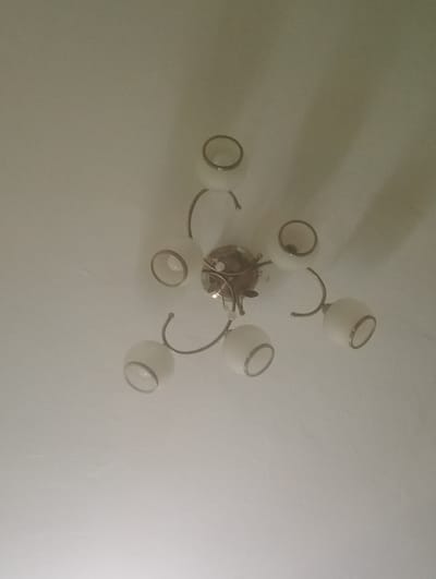 The image shows a chandelier with six white glass shades, each with a metallic rim. The chandelier is mounted on a white ceiling. The overall lighting is dim, and the image quality is poor. The location of this scene in Dybshche, Ukraine is not ascertainable from the image itself.