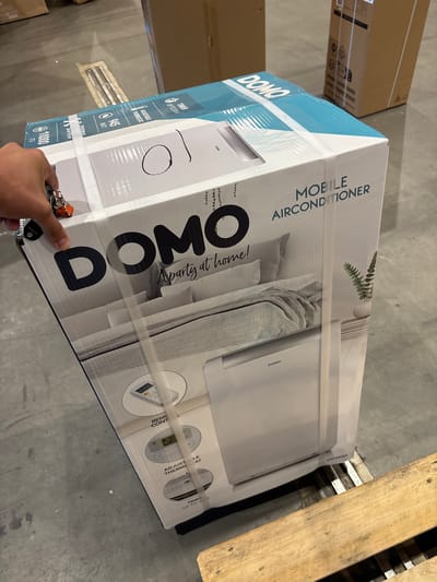 An overhead medium shot shows a white and turquoise cardboard box for a "DOMO MOBILE AIRCONDITIONER" positioned on a wooden pallet. The box is oriented vertically, with the front facing the camera. The brand name "DOMO" is prominently displayed in large black lettering. Below this, text reads "Apartment at home!". The lower portion of the box features an illustration of a bedroom scene with a bed and a white air conditioning unit. Various circular icons detailing features like "REMOTE CONTROL", "ADJUSTABLE THERMOSTAT", and "TIMER UP TO 24H" are visible. A person's hand, holding a keyring with several keys, is reaching from the left side of the frame, partially obscuring the top left corner of the box. The background consists of a concrete floor with other cardboard boxes and industrial elements, suggesting a warehouse or storage environment. The scene appears to be indoors, with artificial lighting. The presence of the wooden pallet indicates the item is likely being stored or prepared for transport.