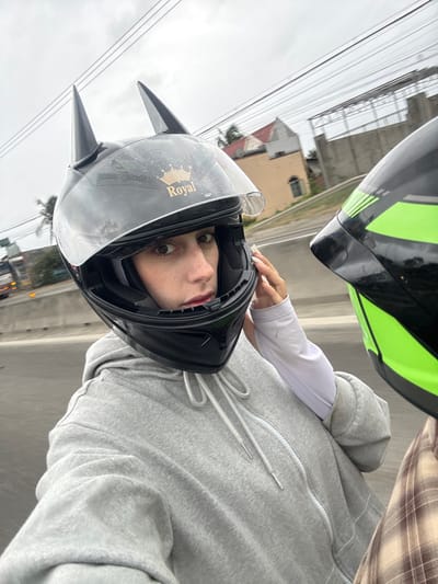 A person wearing a helmet with cat ears, labeled "Royal", faces the camera. They wear a gray hoodie and a white sleeve underneath. Another helmet, green and black, is visible above them. The background includes buildings, power lines, and a cloudy sky. The perspective is tilted, suggesting the photo was taken while in motion or at an angle.