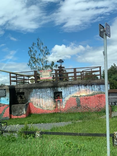 The image shows a concrete fortification, likely a bunker, with a large cannon barrel protruding from the left side. The bunker is painted with a mural depicting a field of red flowers and a figure standing in them, under a sky with clouds. A sign with Russian text "Беларусь помнит" (Belarus Remembers) is attached to the railing on top of the bunker. There is a wooden fence in the foreground, with green grass and some gravel paths. In the background, there are trees and a signpost with a number "13" and other directional signs. The setting appears to be an outdoor historical memorial or museum in Цімкавіцкі сельскі Савет, Belarus.