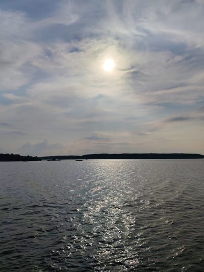 This is a vertical, eye-level, full shot of a body of water with a treeline and sky. The sun is visible in the upper center of the image, partially obscured by wispy clouds. The water is dark and reflects the sunlight in a shimmering path extending towards the viewer. A dark, silhouetted treeline stretches across the horizon in the middle ground, with a few small islands or protrusions visible in the water. The sky is a mix of blue and gray clouds, with some areas of brighter sky near the sun. The location is identified as Bessolovo, Russia. There are no people or animals visible in the image. The environment is natural and appears to be a lake or large river. The overall mood is serene and tranquil.