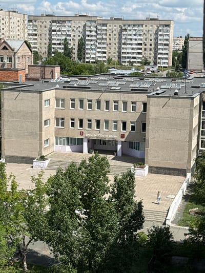 This is a high-angle, eye-level, medium shot of a school building in Novotroitsk, Russia, on a sunny day. The school, named "ШКОЛА ИМ 350 ЛЕТИЯ МОСКВЫ" (School Named After the 350th Anniversary of Moscow), is a multi-story concrete structure with numerous windows. The front of the school features a prominent entrance with stairs and a paved courtyard. A single woman in light-colored clothing is walking away from the school down the stairs. Trees with green foliage are scattered around the school grounds and in the background, obscuring some of the view. In the distance, several large, identical apartment buildings dominate the skyline, a common feature of Soviet-era urban planning. The sky is partly cloudy.