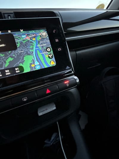 The car's infotainment system displays a navigation map with a route highlighted in blue, showing a distance of 61 km and an estimated time of 54 minutes. The map includes details of roads, green areas, and points of interest. The center console features buttons, one with a red lock symbol illuminated and another with a red triangle for hazard lights. A small white object, possibly a USB drive, is visible in a storage compartment.
