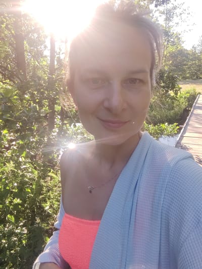 A woman is taking a selfie outdoors near a wooden boardwalk, with lush greenery and trees in the background. The sun is bright, creating a lens flare effect. She is wearing a coral-colored bikini top and a light blue waffle-knit robe. A thin silver necklace with a small pendant is visible around her neck. The scene appears to be set in a natural, possibly rural, environment. The location is identified as Бярэзінскі сельскі Савет, Belarus.