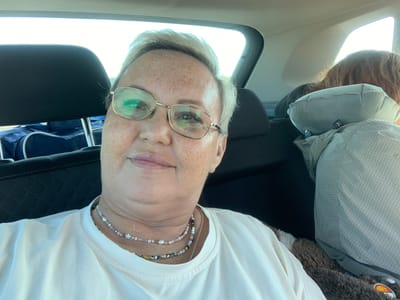 In this close-up, selfie-style photograph taken inside a vehicle, a middle-aged woman with short, blonde hair and fair skin adorned with freckles is the central subject. She is wearing glasses with gold-colored frames, a light-colored t-shirt, and two beaded necklaces. Her gaze is directed towards the camera, and she has a slight smile. The background reveals the interior of a car, with a headrest behind her and a window to her right. Partially visible in the background are luggage and another passenger with reddish-brown hair, whose face is obscured. The lighting suggests it is daytime, possibly a sunny day, casting reflections on the woman's glasses. The scene appears to be a casual moment during a car journey, with no specific indicators of the city of Старое Русское Маматкозино, Russia, other than the context provided.