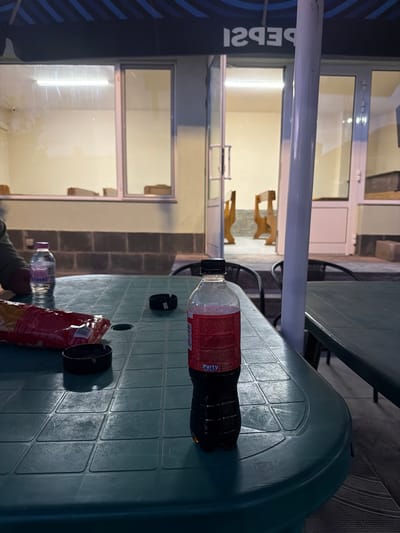 The image depicts an outdoor scene with a green plastic table in the foreground, likely in Byala Reka, Bulgaria. The table has a bottle of Coca-Cola, a bottle of water, a bag of chips, and two black ashtrays. In the background, there's a building with a white door and window frames, showing an interior with chairs. A Pepsi advertisement banner is partially visible on the left side of the frame, attached to a metal pole. The environment suggests a casual outdoor setting, perhaps a patio or terrace adjacent to a building. No people are fully visible, though a partial view of a person's arm can be seen near the bottom of the frame.