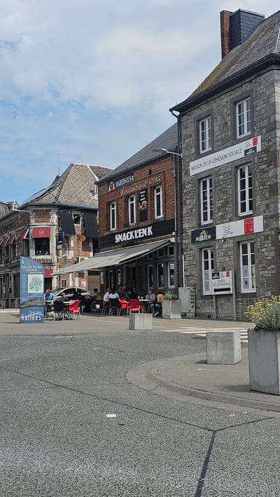 The image captures a view of a street scene in Philippeville, Belgium, featuring several buildings and outdoor seating areas. On the left, a building with awnings displays a sign for "THEY HOUSE" and "Pays des Vallées - The Land of Valleys" with a map. Adjacent to it, a brick building houses "MAREDSOUS RESTAURANT ADETAGE" and "SNACK L'EN K" with outdoor tables and chairs occupied by patrons. To the right, a stone building displays "MAISON DE LA COHESION SOCIALE," "MIRESEM," and a directional sign for "ENTREE RUE DE NAMUR." Several concrete planters with greenery are visible, and the street surface is paved asphalt with concrete curbs. The sky is partly cloudy.