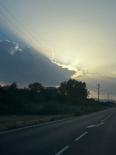 The image depicts a rural roadside scene with a paved asphalt road stretching into the distance, marked with white lines and an arrow indicating a turn. To the left of the road is a dense line of dark green vegetation, likely trees and bushes, with a rough grassy embankment along the roadside. In the background, silhouetted against a bright sky, are more trees and hills. The sky is a dramatic mix of dark clouds and bright sunlight, with rays of light breaking through, creating a strong contrast. Overhead, power lines stretch across the frame, supported by utility poles visible along the right side of the road. The lighting suggests it is either sunrise or sunset, casting a warm, golden hue over the scene. There are no people or vehicles immediately visible. The overall impression is one of a natural landscape adjacent to a man-made road, illuminated by dramatic atmospheric conditions.