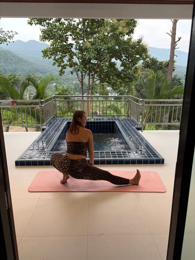 In Ban Kao Dua, Thailand, a woman wearing a leopard-print athletic set is performing a yoga pose on a pink mat. She is positioned on an outdoor tiled patio, facing away from the camera. Behind her is a rectangular swimming pool with blue tile accents, and beyond that, a lush, green landscape with rolling hills and trees. A metal railing runs along the edge of the patio, overlooking the scenic view. The scene is framed by dark vertical structures on either side, suggesting an entrance or doorway.