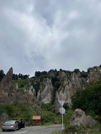 The image displays a segment of the Goris Rock Forest in Armenia, characterized by a steep, heavily vegetated hillside featuring numerous naturally eroded, towering rock spires and columns, ranging in color from light grey to reddish-brown. The sky is uniformly overcast with light grey clouds.

In the lower left, a silver four-door sedan, bearing Armenian license plate '13 SP 033', is parked on an unpaved gravel road. The vehicle's front driver's side door is open. A single individual, dressed in dark clothing, stands beside the open car door, facing inward towards the vehicle's interior. To the right of the parked vehicle, a small wooden structure with a reddish-brown tiled roof and open sides is visible behind a low wooden fence. Further to the right along the road, a large wooden cable spool rests on the ground. A white metal pole supporting two circular grey road signs, the upper sign being an inverted triangle, stands in the mid-ground. A prominent, rounded boulder, partially covered with green vegetation and small flowering plants, occupies the lower right foreground. Dense green foliage, including tall coniferous trees at higher elevations, covers the mountainous terrain. A dark, multi-faceted rock formation or entrance is partially visible on the far left, positioned behind the parked car.