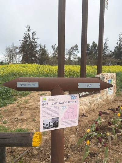 A rusted metal signpost is in a field of yellow flowers. Two signs point in opposite directions, reading "Le'eri Forest" and "Le'Bari HaYishar (Nahabir)". Attached to the post is a paper sign with a map and historical information in Hebrew, titled "The First Water Line to the Negev - 1947". The post is mounted on a wooden stake painted with yellow at the top. The background features trees and a cloudy sky.