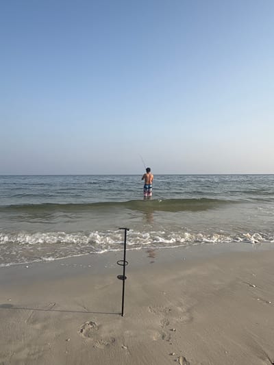 An individual is fishing in the calm waters of the Arabian Gulf near the city of Ajman, United Arab Emirates. The person stands waist-deep in the sea, holding a fishing rod that arcs over the water. They are wearing colorful patterned swim shorts. The foreground of the image shows a sandy beach with footprints and a black fishing rod holder planted in the sand. Gentle waves lap at the shore, creating a white foamy edge. The sky is clear and blue, with a hazy horizon where the sea meets the sky. The overall scene depicts a peaceful coastal activity.