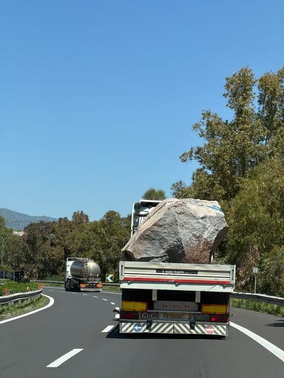 The image captures a bright, sunny daytime scene on a highway near Sant'Alessio Siculo, Italy. The photograph is taken from a moving vehicle, looking forward.

In the foreground, a large flatbed truck is prominently featured, transporting an enormous, irregularly shaped grey rock or boulder. The rock dominates the truck's bed, secured with visible blue strapping on its upper surface. Behind this leading truck, a smaller tanker truck follows on the same lane. Both vehicles are on a paved highway with clear white lane markings. Trees and lush greenery line the sides of the road, and distant mountains or hills are visible under a clear blue sky. Metal guardrails run along the shoulder of the highway.

Notable details include:
- **Visible Text:** On the rear of the leading truck's trailer, the company name "L AGRO MARMI s.r.l." is visible, suggesting a stone or marble transportation business. License plate numbers "FN 587AV" and "AE 272DB" are also present. Various road safety markings include a circled "80" km/h speed limit sign, a "70" km/h sign on a yellow background, diagonal black and white reflective stripes, and a white arrow in a blue circle. A red circular sign with a diagonal line (prohibitory) is also visible.
- **Roadside Signs:** A yellow square sign with a black downward arrow is present on the right side of the highway.
- **Weather and Time:** The clear blue sky and bright illumination indicate good weather conditions during daylight hours.

The image depicts the logistical challenge and scale of transporting large natural rock formations on public roads.