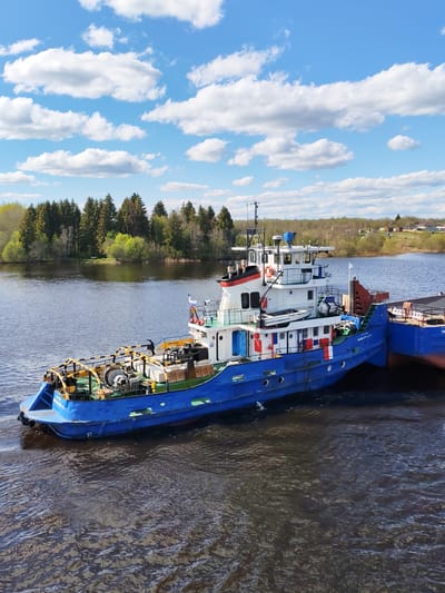 The image depicts a blue and white tugboat on a body of water, likely a river or large lake, near Kochino, Russia. The tugboat is positioned close to a barge, which is partially visible on the right side of the frame. The tugboat has various equipment on its deck, including winches and cables. The landscape in the background features a line of trees and a clear blue sky with scattered cumulus clouds. There is no visible street name, and the only visible text says "SHAPOSHNIKOV-111".