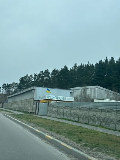 The image shows a building located in Yurivka, Ukraine at coordinates 50.328, 30.375. The building is a long, rectangular structure with a gray exterior. It is surrounded by a gray concrete fence with decorative designs. A Ukrainian flag is mounted on the building. The word "OXOPONA" is displayed prominently on the building. The building is situated in a residential area, surrounded by a line of trees. There is a paved road in the foreground with a sidewalk on the left, and green grass on the right. No people are visible. The weather is overcast. The scene is taken from a street, looking towards the building.