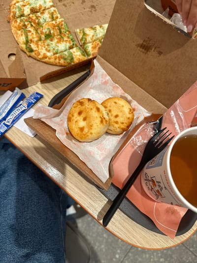 The image shows a table in a food establishment, likely a fast-food restaurant, in the city of Sverdlovskiy, Russia. On the table, there is a pizza with green sauce and cheese, cut into squares, in a cardboard box. Next to the pizza are two small, round, golden-brown baked goods, possibly buns or rolls, served on a piece of patterned paper. There are also two small blue packets, likely condiments or seasonings, and a black plastic fork. In the foreground, a white paper cup filled with a light brown liquid, possibly tea, sits on an orange paper napkin. The table surface is made of light-colored wood. The overall scene suggests a casual meal setting.