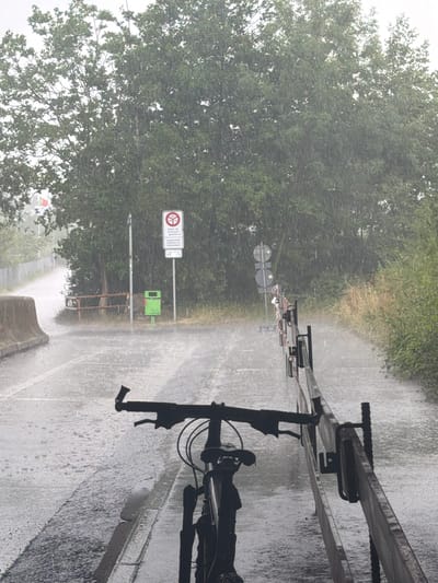 In Wallisellen, Switzerland, during a heavy rain shower, a bicycle stands on a wet asphalt road. The bicycle is in the foreground, silhouetted against the rain-streaked environment. Behind it, a wooden barrier runs along the right side of the road. The road itself is covered in puddles, with rain visibly falling. In the background, a dense green treeline borders the road, and a traffic sign is visible, indicating a prohibition. A green trash bin is positioned near the sign. The overall atmosphere is damp and overcast due to the rain.