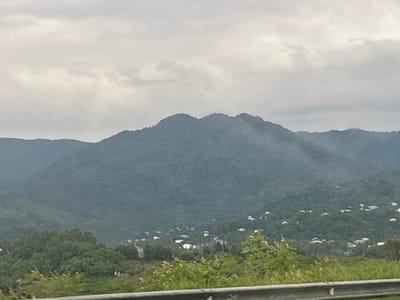 The image shows a misty mountain landscape with scattered houses nestled on the slopes. The foreground features a metal guardrail and green vegetation. The sky is overcast with clouds. The scene appears to be in a rural or semi-rural area, with a sense of distance and depth created by the layered mountains. The houses, primarily white, are small and appear to be residential buildings. The overall atmosphere is subdued due to the weather conditions.