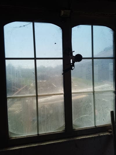 The image shows a close-up view of a dusty, arched window with multiple panes. The window looks out onto an outdoor scene that is hazy and indistinct, with hints of greenery and structures. A dark object, possibly a piece of machinery with a circular serrated component, is hanging from the window frame on the right side of the image. The interior foreground is dark and appears to be a concrete ledge. The scene is lit by natural light filtering through the window. The location is identified as Nicorești, Romania.