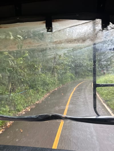 The image displays a rain-soaked asphalt road curving to the right, viewed from inside a vehicle through a translucent, wet plastic or vinyl window panel. The panel's upper edge is secured by multiple dark fasteners to a dark overhead structure. A solid yellow line marks the center of the wet road surface. Dense green foliage, comprising trees and bushes, flanks both sides of the roadway. On the left, a shoulder with scattered brown leaves is visible, along with a thin blue line or wire positioned horizontally within the vegetation. The right side of the road features a grassy verge leading to similar dense foliage, partially obscured by a dark, vertical structural element of the vehicle. No individuals or other vehicles are present in the scene, which is located in the city of Ban Paem Bok, Thailand.