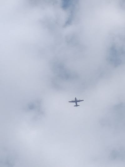 An airplane flies in a cloudy sky. The aircraft is silhouetted against the gray and white clouds, viewed from below, showing the plane's underside. Its wings are extended, and it appears to be a four-engine turboprop, possibly a military transport. The clouds fill the majority of the frame, offering a stark contrast against the dark silhouette of the airplane.