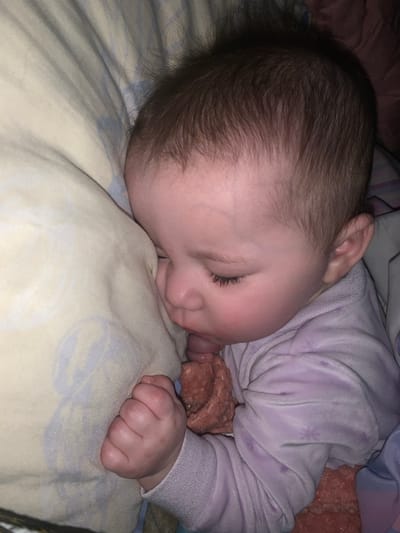 The image displays a sleeping infant resting on its left side, likely in a bed. The infant has light skin, dark brown hair, and closed eyes with distinct eyelashes; its cheeks show a rosy hue. It is dressed in a light purple, long-sleeved garment, appearing to be made of velvet or velour, with faint patterns. The infant's face is partially nestled into a pale yellow or beige pillow that features subtle light blue designs. A crumpled, reddish-brown fabric item is partially held within the infant's mouth and extends beneath its right arm. The infant's right hand is clenched into a fist near its chest. In the background, additional bedding, possibly a blue and white striped or patterned fabric, is visible. The specific location within the city of Lancaster, United States of America, cannot be determined from the visual information provided.