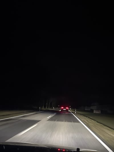 A nighttime view shows a road stretching into the distance, illuminated by headlights. The asphalt gleams with moisture, reflecting the light. The left side is dominated by darkness, while the right is defined by a white line marking the road's edge. Red taillights of several vehicles are visible further ahead, indicating traffic. The composition is heavily reliant on the contrast between the dark background and the illuminated road.
