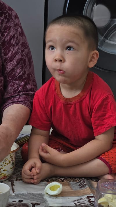A young boy, likely East Asian, is seated on the floor, cross-legged, in a domestic indoor setting. He is wearing a red t-shirt and red shorts with a polka dot pattern. His gaze is directed upwards and to his left, with his lips slightly pursed. Beside him, on his left, is an adult's arm and shoulder, draped in a dark purple, patterned fabric. On the floor in front of the boy are several items: a half-peeled hard-boiled egg, a white ceramic bowl with a floral design, and a transparent plastic container filled with chopped food, possibly more egg. The surface beneath them is a patterned rug or tablecloth. In the background, to the right, a portion of a large, metallic appliance, likely a washing machine, is visible. The lighting appears to be natural and diffused. The scene is likely within a residential home in Mary City, Turkmenistan, given the cultural context implied by the context of the image.