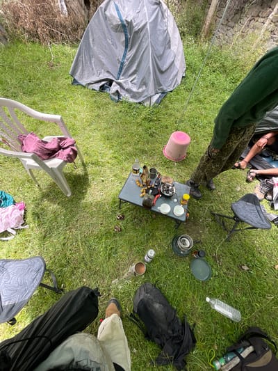 A high-angle, wide shot shows a campsite in Panamik, India. The scene is set on a grassy area with a large, grey tent partially visible in the background. A white plastic chair holds a folded maroon garment. A low, foldable table is laden with camping cooking equipment, including a stove, pots, pans, cups, and food items such as small yellow fruits and bottles. Two folding chairs are positioned around the table, one with a patterned seat. A person in green and leopard-print clothing stands near the table, and another person's legs are visible in the lower right, wearing sandals. Various camping gear, including backpacks and sleeping bags, are scattered on the ground, along with a pink bucket and a water bottle. The overall impression is of a casual outdoor gathering or camping setup.