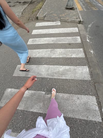 This is a high-angle, first-person perspective shot of two individuals crossing a pedestrian crosswalk. The scene is set on a paved road with clearly marked white stripes for the crosswalk. To the left of the crosswalk, a sidewalk made of interlocking grey pavers is visible, with some sparse green vegetation growing through the cracks. A metal street lamp post is also present on the left side of the frame.
One person, closer to the viewer, is wearing light blue pants and black flip-flops. Their arm is extended to the right, with a black bracelet on their wrist. The other person, further into the crosswalk, is wearing a lilac-colored skirt or pants and white flip-flops with black polka dots. They are also wearing a white garment, possibly a shirt or dress, draped over their shoulder.
The lighting suggests it is daytime, with shadows cast across the crosswalk. The weather appears to be overcast, as there are no harsh shadows. The setting of Derbent, Russia, is provided as context but not visually identifiable from the image itself. No visible text is present in the media file. The activity is simply crossing the street, and the overall mood is casual.