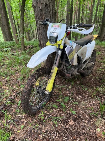 A white and yellow dirt bike lies on its side in a forest, possibly in Gruzinskii, Russia. The bike is covered in mud, suggesting recent use. The surrounding environment consists of trees, grass, and leaf litter. There are no visible people in the image. The location is likely a wooded area near or in Gruzinskii. The interaction is implied: the bike was being ridden in the area and has fallen over.