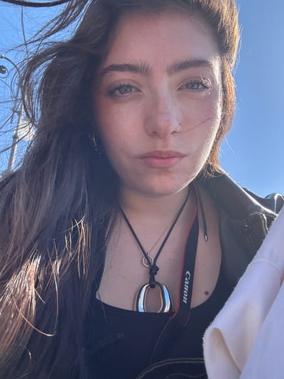A low-angle selfie of a young woman with brown hair, light skin, and freckles. She has a nose ring in her left nostril and distinct eyebrows. She is wearing a black tank top, a dark green jacket, and a white hooded sweatshirt. A black camera strap with the word "Canon" in white lettering is around her neck, along with a silver pendant on a black cord and a small, delicate necklace. The sky is a clear blue. The setting appears to be outdoors, possibly in Sorrento, Italy, judging by the type of light, the plants visible at the bottom of the image, and the general aesthetic.