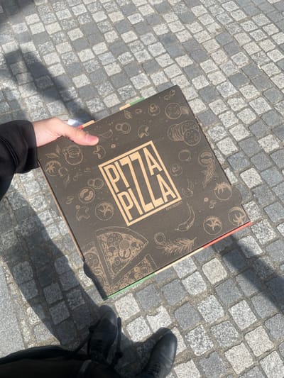 A first-person, downward-angled view captures an individual holding a dark brown pizza box outdoors in Montigny-le-Bretonneux, France. The visible right hand, covered by a black sleeve, grips the box. The box's lid features "PIZZA PIZZA" in light brown lettering, accompanied by outline illustrations of various pizza ingredients such as mushrooms, salami, tomatoes, and herbs, and the text "Bon appétit." Green and red colored tabs or stripes are visible on the box's side edges, suggestive of the Italian flag. The individual's lower legs and feet, wearing black shoes, are partially visible at the bottom of the frame. The ground consists of grey cobblestone paving. Direct sunlight casts distinct, elongated shadows of the individual's arm and legs onto the pavement.