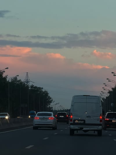 This image depicts a view from a vehicle traveling on a multi-lane highway at dusk, likely on the outskirts of Krasnaya Pakhra, Russia, given the presence of Russian license plates and the general landscape. The sky is a soft gradient of pale blue and pink, with scattered clouds illuminated by the setting sun. Along the right side of the highway, a row of tall streetlights with multiple lamps lines the road, interspersed with trees. A power transmission tower is visible in the distance, silhouetted against the sky behind the trees. Several vehicles are on the highway, including a white van in the foreground, a white sedan in the left lane with illuminated taillights, and other vehicles further ahead, some with headlights or taillights visible. The road surface is asphalt with white lane markings. The overall atmosphere is serene and indicative of evening traffic.