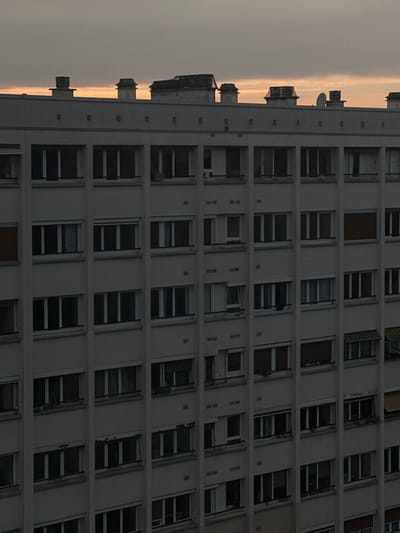 An exterior, medium-angle photograph captures the facade of a multi-story residential building in Maisons-Alfort, France, under low-light conditions, indicative of either dawn or dusk. The structure is a light-colored concrete apartment block, characterized by a uniform grid of rectangular windows and recessed balconies across its visible eight to nine floors.
The windows display various conditions: some are fully closed, others are partially open via a tilt-in mechanism, and many are covered by closed or partially closed roller shutters. Internal curtains are visible in several windows. Distinct objects include a reddish-brown plant pot situated on a balcony railing on the second floor from the top, second column from the left. A red fabric item is draped over a railing on a balcony on the same floor, fifth column from the left. A dark garment hangs from a window on the fifth floor from the top, second column from the right. Several lower-level windows feature partially extended striped awnings.
The flat rooftop hosts multiple ventilation shafts and chimneys of varying designs, including one small satellite dish positioned on the right side. A narrow band of orange-pink light is visible along the horizon, directly above the rooftop, contrasting with the otherwise overcast gray sky. No individuals or dynamic interactions are visible within the frame.