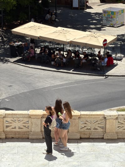 An outdoor cafe in Cagliari, Italy, is depicted from an elevated perspective on a sunny day. In the foreground, three young women stand on a marble-tiled terrace behind a stone balustrade with decorative geometric cutouts. The woman on the left wears dark trousers, a black top, and a light-colored backpack, facing left with her arms crossed. The middle woman wears a pink top and denim shorts, holding a small green object and a dark bag. The woman on the right also wears denim shorts and a light pink top, holding a dark bag.
Behind the balustrade, the cafe features numerous tables and chairs set up under large, beige awnings. People are seated at the tables, engaged in conversation and eating or drinking. Some of the patrons are visible in the midground. A server in a white shirt and black apron stands near the counter area on the left side of the cafe. Further in the background, trees provide shade, and a promotional cube with colorful graphics stands on the pavement, indicating an event or exhibition. The cafe appears to be located on a street or public square, with a road visible beyond the seating area.
