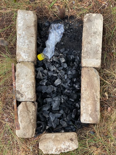 An overhead view shows a makeshift fire pit constructed from concrete blocks, filled with charcoal. A crumpled plastic bottle with a yellow cap lies on top of the charcoal. The fire pit is surrounded by dry grass and pine needles, indicative of an outdoor, possibly wooded, environment. There are no people or any overt actions taking place within the frame. The scene appears to be in Leninsky, Russia, based on the provided location.