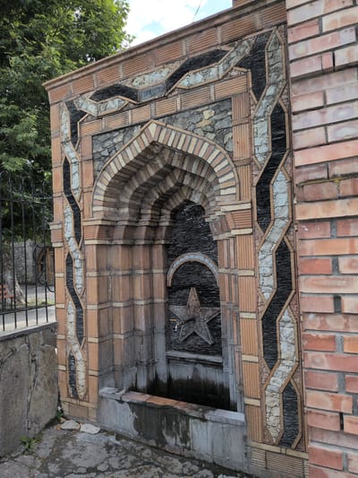 The image shows a decorative water fountain built into a brick wall in the city of Şəki, Azerbaijan. The fountain is constructed with a variety of materials, including reddish-brown bricks, grey stone, and black slate. It features an arched niche that houses a star-shaped relief with a faucet. The surrounding area is paved with stone and has some vegetation. A black metal fence is visible to the left of the fountain, and a large tree with green leaves is in the background. The overall impression is of a historical and artisanal water feature integrated into the architecture of the city.