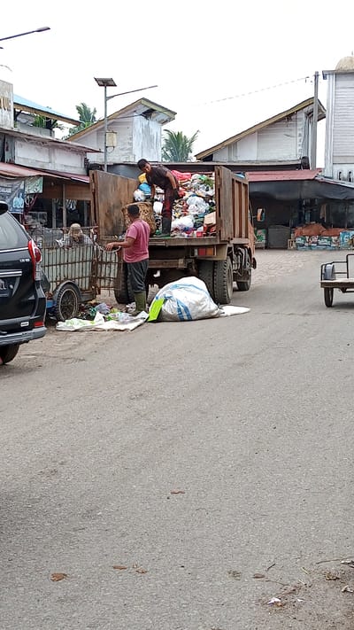 In Puloara, Indonesia, a scene unfolds on a paved street, suggesting an area of urban activity. Two individuals are engaged in waste management. One person, wearing a dark t-shirt and dark pants with green rubber boots, is positioned inside a large, rusty truck filled with an extensive amount of refuse. They appear to be actively sorting or loading the trash. The truck itself is substantial, with visible tires and a weathered appearance.
Beside the truck, another individual, dressed in a pink shirt and dark pants, also wearing green rubber boots, stands on the street. They are near a metal cart with a cage-like structure, also containing waste, and a separate large white and blue plastic bag of trash. Several other smaller pieces of trash and bags are scattered on the ground around the truck and cart, indicating the ongoing process of waste collection and disposal.
In the background, buildings with corrugated metal roofs and concrete walls are visible, suggesting a commercial or residential area. A black SUV is partially visible on the left side of the frame, adding to the sense of an active street scene. The sky is overcast. The overall impression is one of daily work related to waste management in a developing urban environment.