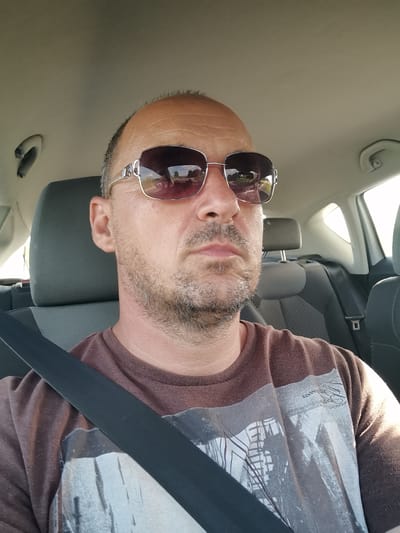 A close-up, medium shot, taken from a high angle, shows a man sitting in the driver's seat of a car. The man is wearing sunglasses and a brown t-shirt with a graphic design. He has short, graying hair and a beard. The man's head is slightly tilted to the right. The interior of the car is visible behind him, including the headrest of the passenger seat. The image was taken in Stefanovo, Bulgaria.