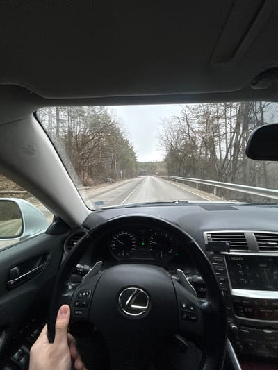 The image presents a driver's perspective from within a Lexus vehicle, looking forward onto a two-lane asphalt road in Sarantsi, Bulgaria. The road, appearing damp, is bordered by bare deciduous trees on both sides, suggesting a winter or early spring season under an overcast sky. A metal guardrail extends along the right side of the road, and a circular road sign is visible on the left in the mid-distance. Inside the vehicle, a light-skinned human hand grips the lower-left section of a black, multi-function steering wheel featuring a silver Lexus emblem and visible paddle shifters. The instrument cluster, displaying two primary gauges, and a portion of the infotainment system with "CLIMATE" and "SOUND" buttons are visible to the right. The driver's side A-pillar is partially visible, showing "SRS CURTAIN AIRBAG" text.
