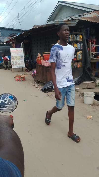 The image depicts a street scene in Agbarho, Nigeria, with a young man walking towards the right of the frame. He is wearing a white t-shirt with blue abstract prints and light blue denim shorts, and black flip-flops. The background shows a row of informal shops with corrugated metal roofs. One shop displays a variety of goods, including what appear to be engine oils and other automotive fluids in containers. To the left, a small stand advertises "OMONICE PROVISION & PEPPER SOUP" with images of food items and a phone number. A few other people are visible in the distance, including a person sitting on a motorcycle and another crouched down. The ground is unpaved and appears to be dusty or sandy. A discarded red and white box, possibly containing food items, lies on the ground. In the foreground, a motorcycle's side mirror is partially visible on the left side of the image. The sky is overcast.